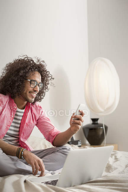 Souriant jeune homme utilisant un téléphone intelligent à l'ordinateur portable sur le lit — Photo de stock