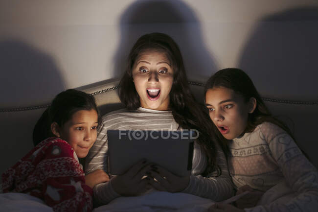 Здивована мати і дочки дивиться фільм на цифровому планшеті в темній спальні — стокове фото