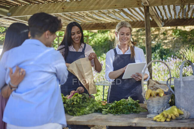 Mujeres con tableta digital ayudando a clientes en el mercado de agricultores - foto de stock