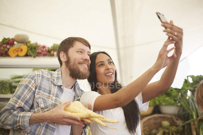 Pareja sonriente tomando selfie en el mercado de agricultores - foto de stock