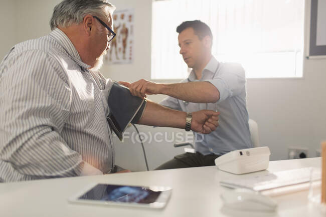 Médico varón que revisa la presión arterial del paciente mayor en el consultorio médico - foto de stock