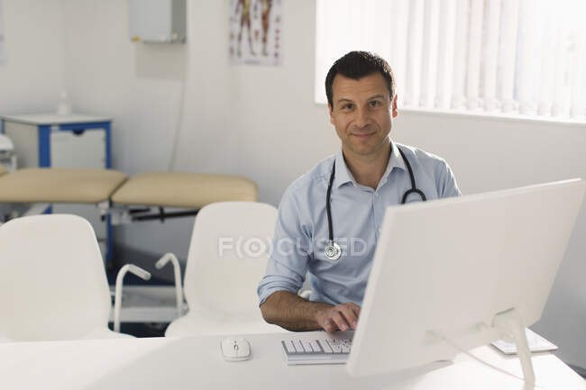Retrato confiado médico varón que trabaja en la computadora en la oficina de médicos - foto de stock
