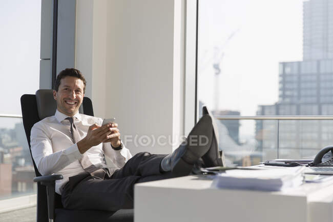 Portrait confident businessman using smart phone with feet up on desk in highrise office — Stock Photo