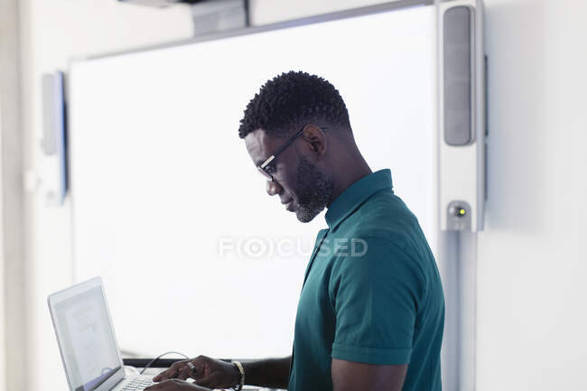 Profesor masculino usando el ordenador portátil junto a la pantalla de proyección en el aula - foto de stock