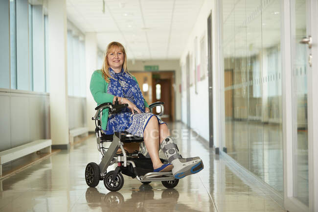 Retrato sorridente mulher vestindo bota médica em cadeira de rodas no corredor — Fotografia de Stock