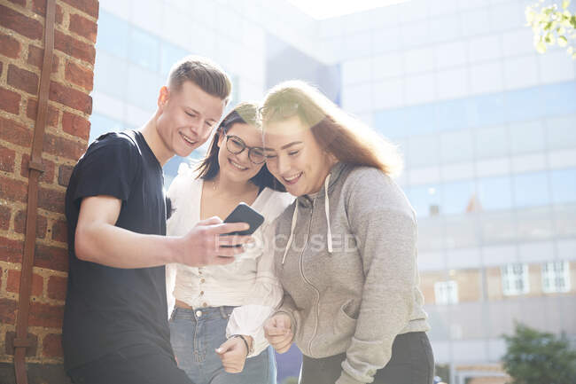 Amigos adolescentes usando un teléfono inteligente fuera del edificio de la escuela soleada - foto de stock