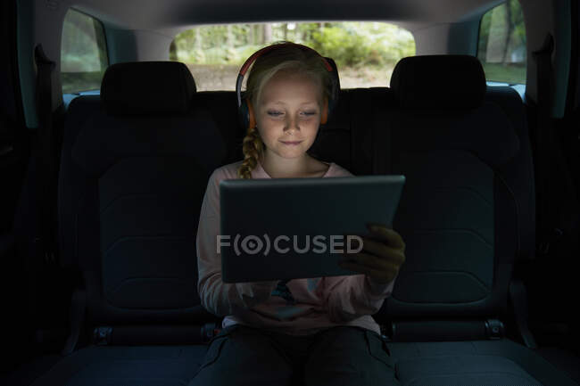 Chica con auriculares y tableta digital en el asiento trasero del coche - foto de stock