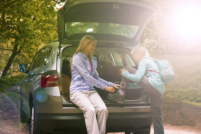 Mother helping daughter tie hiking boots at back of sunny car — Stock Photo