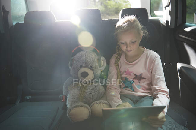 Mädchen mit Teddybär nutzt digitales Tablet auf dem Rücksitz des Autos — Stockfoto