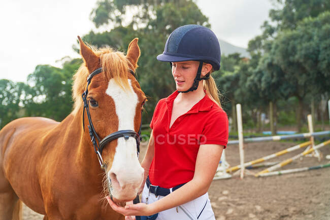 Adolescente en casco ecuestre con caballo en paddock - foto de stock