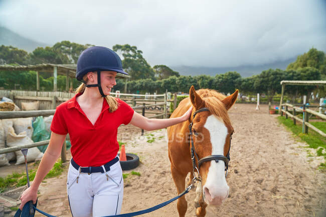 Teenage girl in equestrian helmet leading horse along paddock — Stock Photo