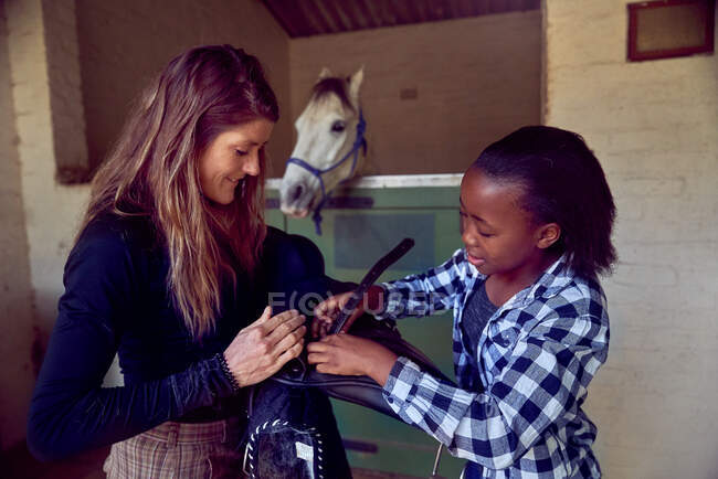Mujer ayudando chica preparar silla de montar en establos de caballo - foto de stock