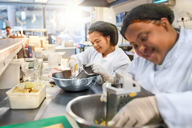 Smiling young women with Down Syndrome grating cheese in cafe kitchen — Stock Photo