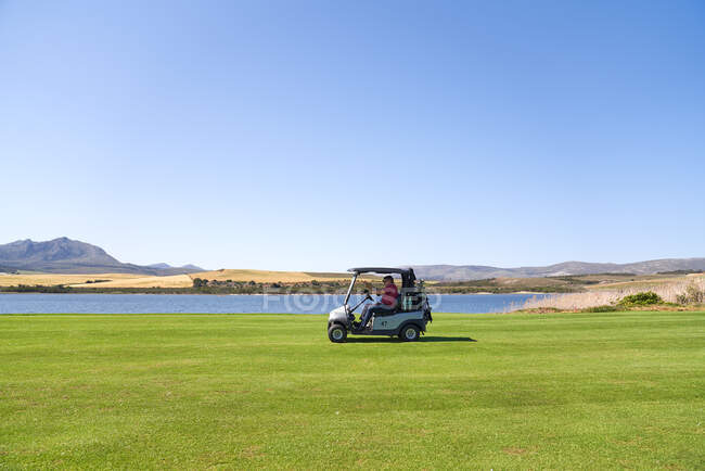 Игрок мужского пола водит гольф-мобиль вдоль озера на солнечном поле для гольфа — стоковое фото