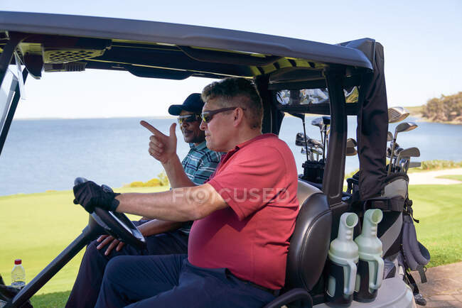 Golfistas masculinos dirigindo carrinho de golfe no campo de golfe à beira do lago — Fotografia de Stock