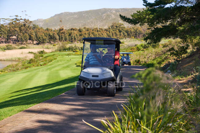 Golfistas masculinos montando no carrinho de golfe no caminho ensolarado do campo de golfe — Fotografia de Stock