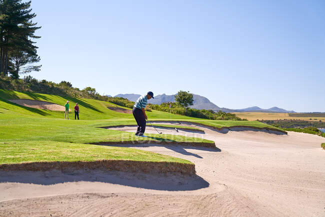 Golfista masculino tomando un tiro por encima de la trampa de arena soleada - foto de stock
