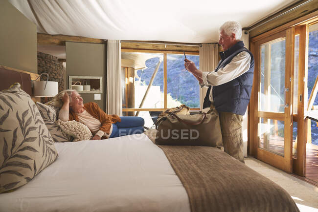 Старший мужчина с фотоаппаратом фотографирует жену на кровати отеля — стоковое фото