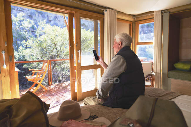 Homem sênior usando telefone inteligente na cama do hotel — Fotografia de Stock