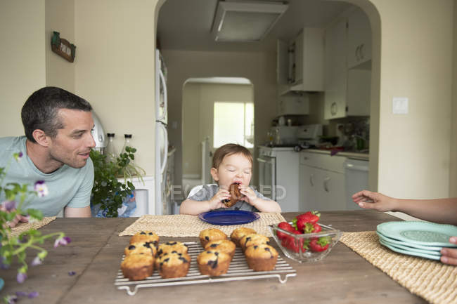 Father watching cute toddler daughter eating muffin at kitchen table — Stock Photo
