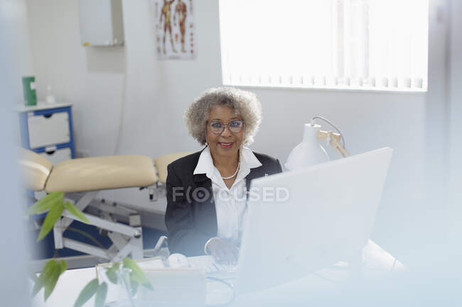 Retrato sonriente, confiado médico senior femenino que trabaja en la computadora en la oficina de médicos - foto de stock