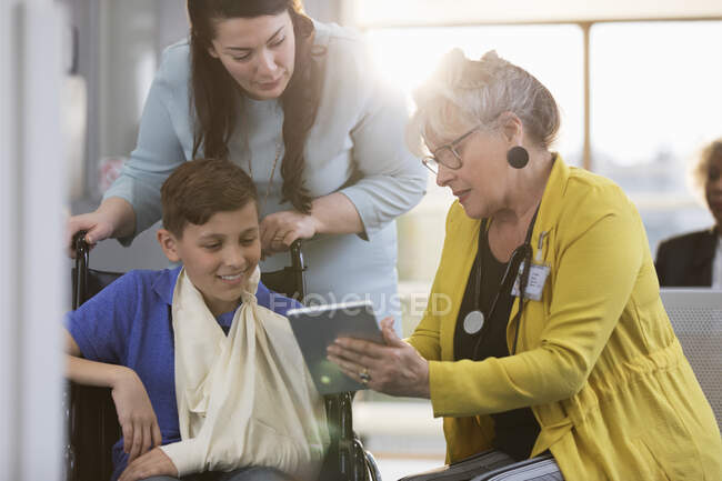 Доктор з цифровим планшетом, що розмовляє з пацієнтом-хлопчиком у інвалідному кріслі та матір'ю у фойє клініки — стокове фото