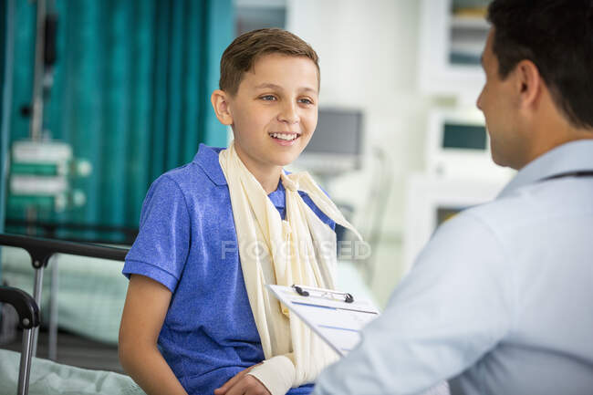 Pediatra do sexo masculino conversando com menino paciente com braço na funda na clínica — Fotografia de Stock