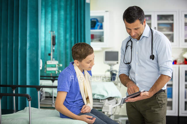 Мужчина педиатр показывает цифровой планшет пациенту с рукой в стропе в больнице — стоковое фото