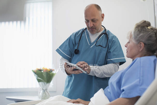 Médico con tableta digital haciendo rondas, hablando con paciente mayor en cama de hospital - foto de stock