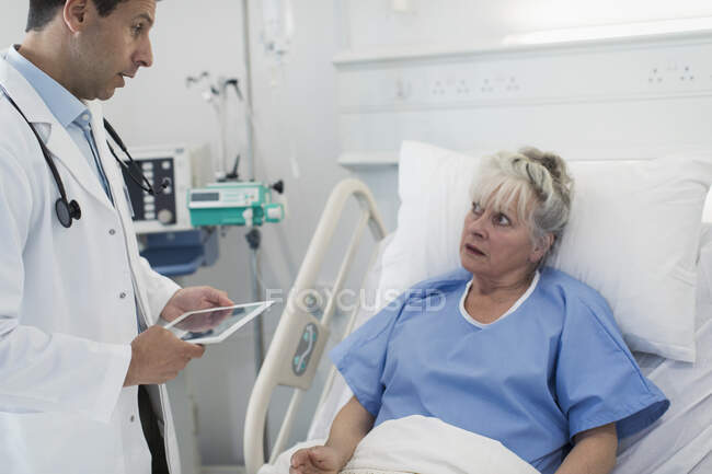 Врач с цифровыми таблетками делает обходы, разговаривает со старшим пациентом в больничной койке — стоковое фото