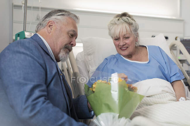 Homme âgé apportant bouquet de fleurs à la femme se reposant dans le lit d'hôpital — Photo de stock