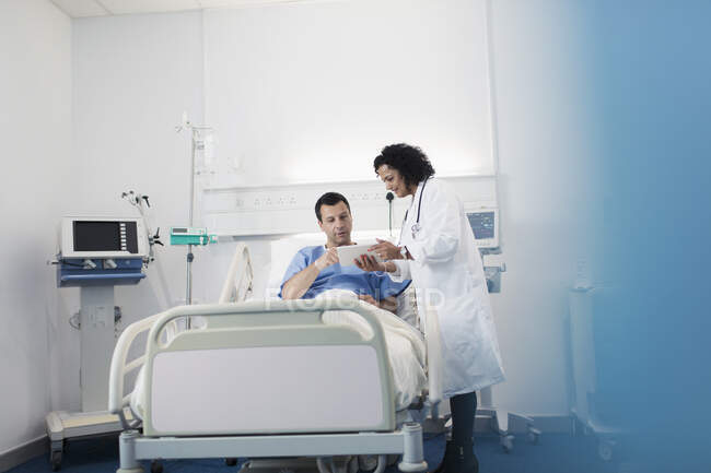 Доктор з цифровим планшетом робить раунди, розмовляючи з пацієнтом, який відпочиває в лікарняному ліжку — стокове фото