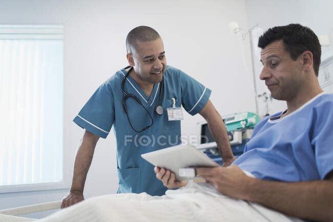 Чоловіча медсестра розмовляє з пацієнтом, використовуючи цифровий планшет у лікарняному ліжку — стокове фото