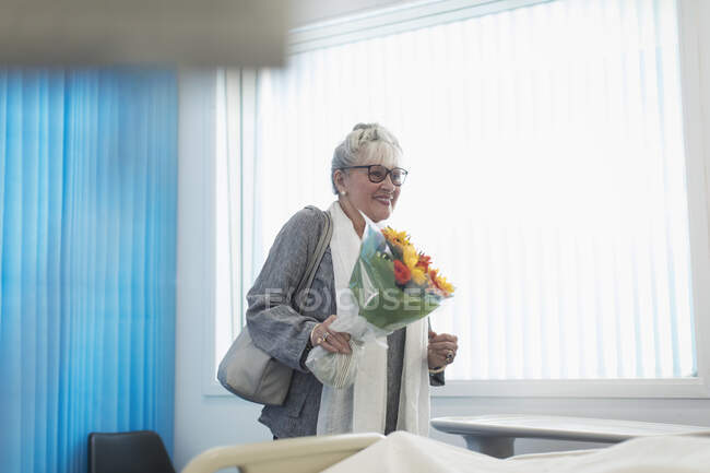Sonriente visitante senior con ramo de flores en la habitación del hospital - foto de stock