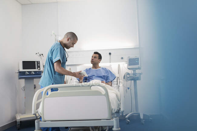 Чоловік лікар з цифровим планшетом робить раунди, розмовляє з пацієнтом у лікарняній кімнаті — стокове фото
