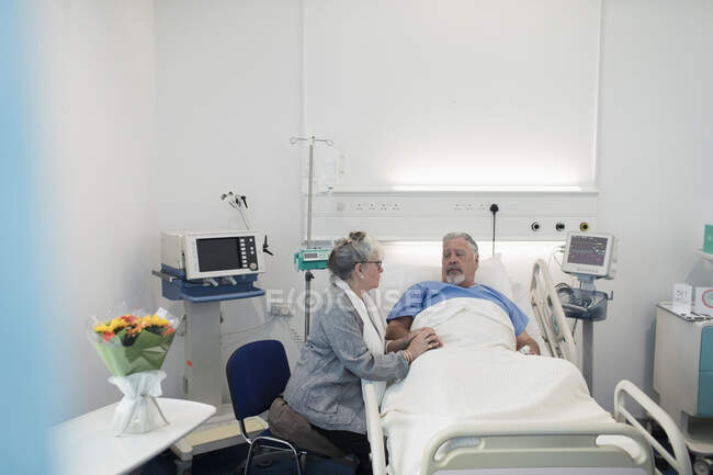 Старша жінка, яка відвідує, заспокоює чоловіка, який відпочиває в лікарняному ліжку — стокове фото