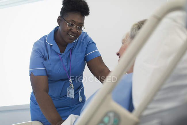 Улыбающаяся, заботливая медсестра разговаривает с пациенткой на больничной койке — стоковое фото