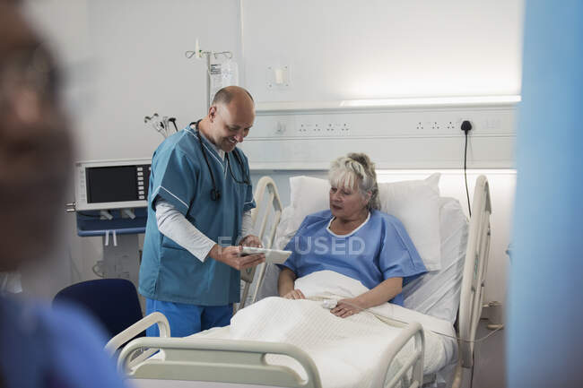 Doctor con tableta digital haciendo rondas, hablando con paciente mayor en la habitación del hospital - foto de stock