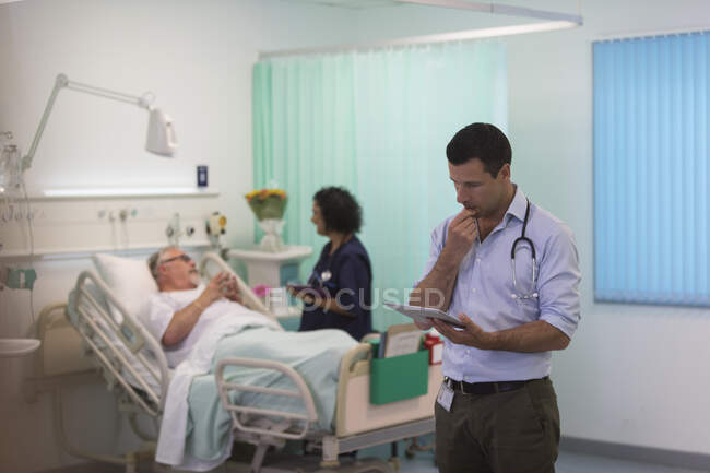 Médico masculino con tableta digital haciendo rondas en la habitación del hospital - foto de stock