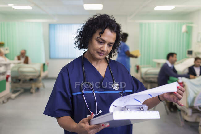 Médico femenino enfocado haciendo rondas, mirando la historia clínica en la sala de hospital - foto de stock