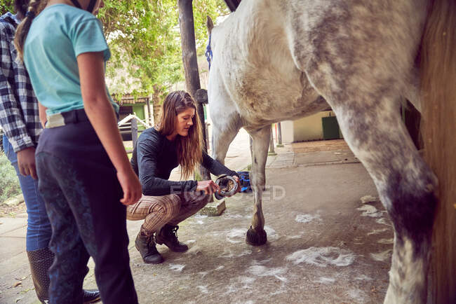 Woman teaching girls how to clean horse hoof outside stables — Stock Photo