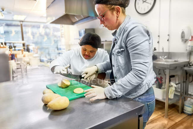 Chef y mujer joven con síndrome de Down cortando papas en la cafetería - foto de stock