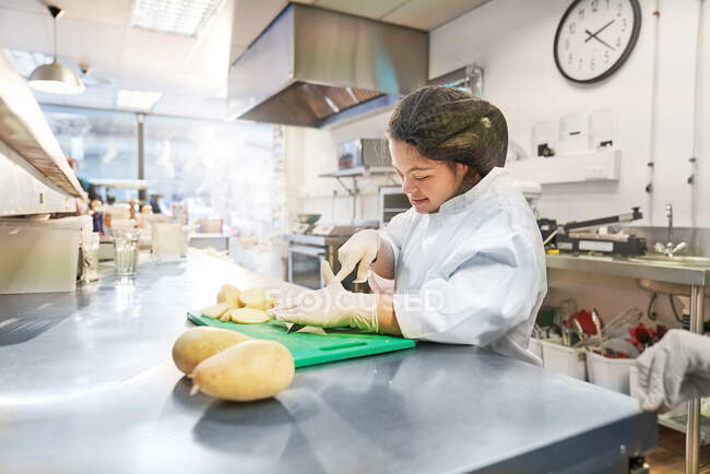 Mujer joven con síndrome de Down cortar patatas en la cocina cafetería - foto de stock
