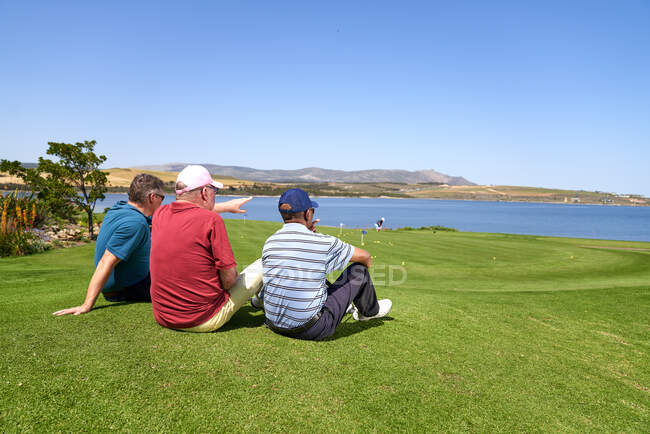 Чоловіки гольфи розслабляються, дивлячись на озеро з сонячного поля для гольфу — стокове фото