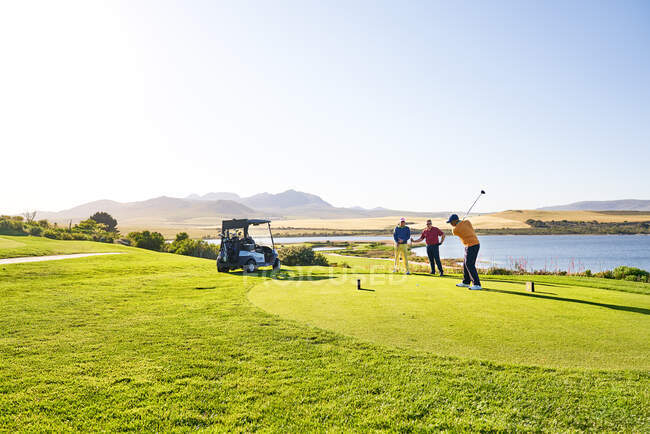 Male golfer teeing off at sunny lakeside tee box — Stock Photo