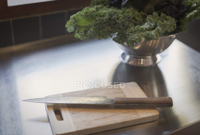 Still life knife on cutting board next to kale in colander — Stock Photo