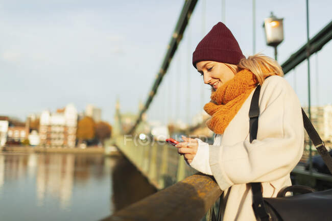 Young woman in stocking cap and scarf using smart phone on autumn bridge — Stock Photo