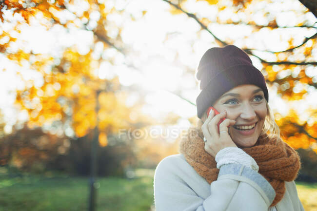 Mujer joven en gorra de siembra y bufanda hablando por teléfono inteligente en soleado parque de otoño. - foto de stock
