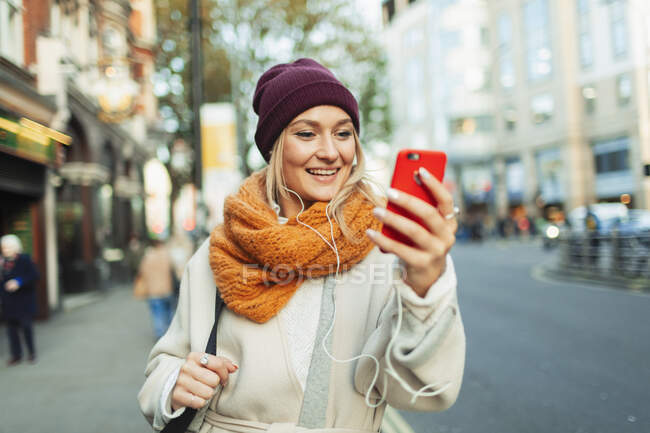 Mujer joven con audífonos y video chat de teléfono inteligente en la calle de otoño - foto de stock