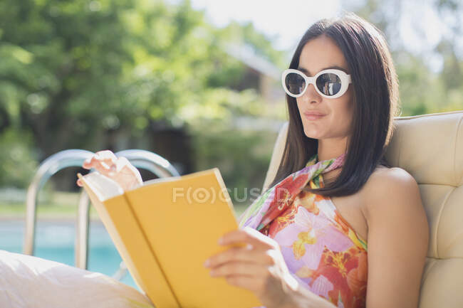 Frau mit Sonnenbrille liest Buch am sonnigen Sommerpool — Stockfoto